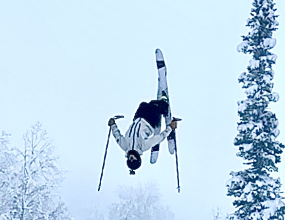 Skier in a white jacket performing a backflip against a pale winter sky with snow-covered trees