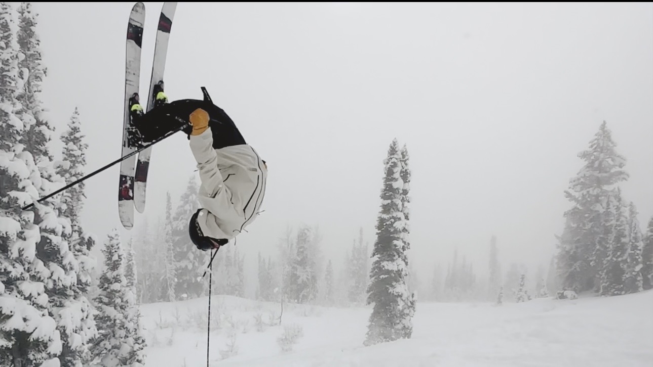 Oskar Lighthiser inverted mid-backflip in a whiteout, skis crossed above a foggy line of snow-covered trees