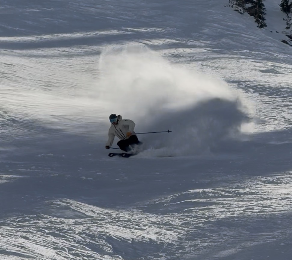 Oskar Lighthiser making a deep powder turn on a wide snowy slope, spraying snow behind him