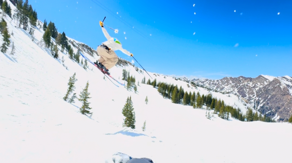 Oskar Lighthiser launching off a snowy slope into a bluebird sky above a tree-lined run